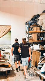 Zwei Personen mit Arbeitskleidung gehen in einem Lagerraum. Im Hintergrund sind Regale mit Materialien zu sehen.